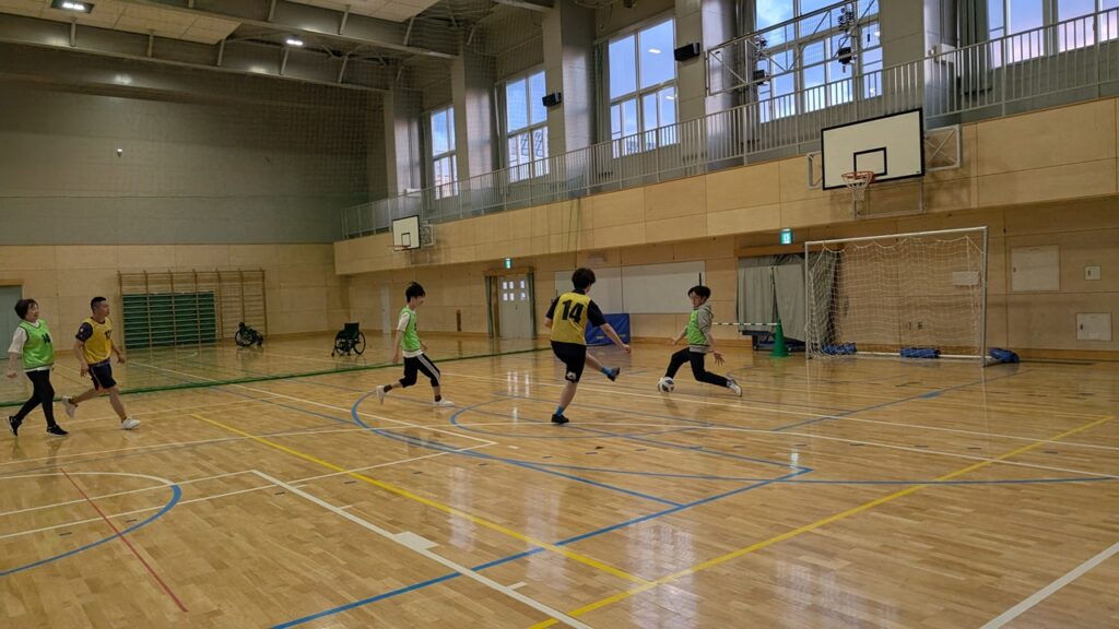 Children playing indoor soccer in a gym; a player in yellow kicks toward the goal while teammates watch.