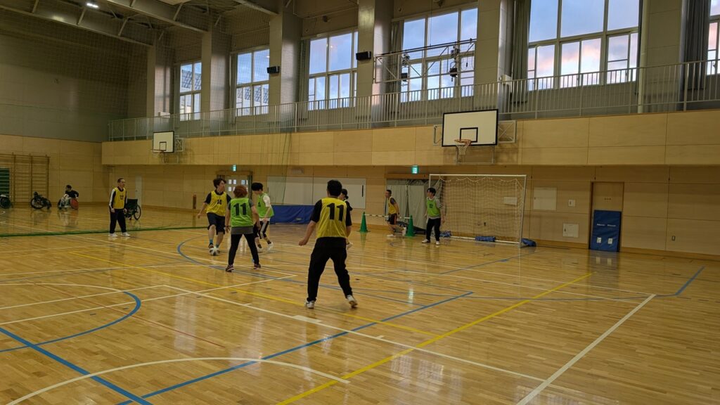 Indoor gym with players in yellow and green pinnies playing soccer; a goal net is visible in the background.