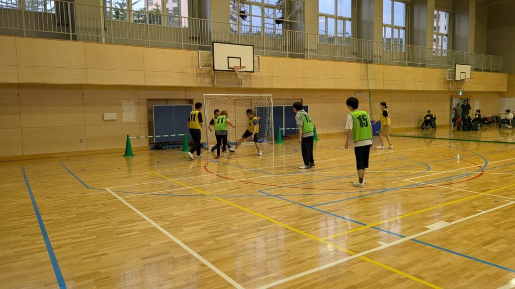 Indoor gym with kids in green pinnies playing small-goal soccer near a goalpost.